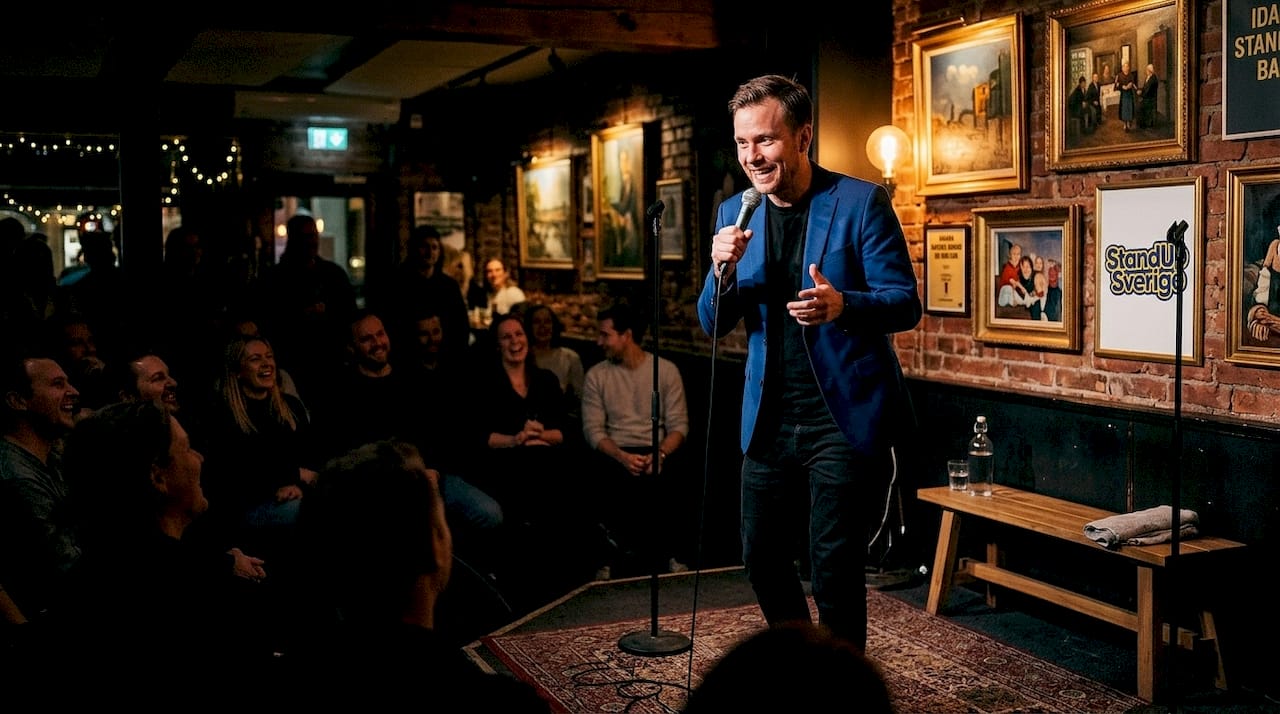 En ståuppkomiker levererar skämt inför en skrattande publik på scenen.