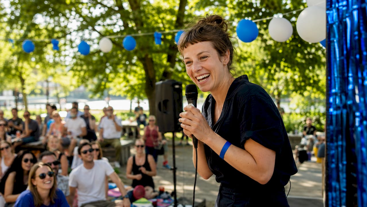 1782014699881_Kvinnlig-komiker-pa-scen-under-standupshow En kvinnlig ståuppkomiker uppträder på scen inför publiken.